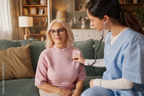 A nurse checks the heartbeat of an elderly woman at home. The nurse uses a stethoscope while sitting on a couch. A cozy living room is visible in the background.