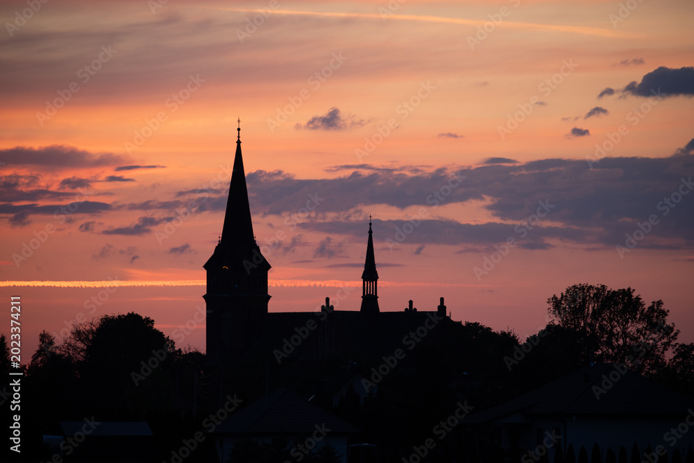 Naklejka premium A picturesque sunset over a quiet town, with the silhouette of a church silhouetted against a vibrant orange and pink sky.