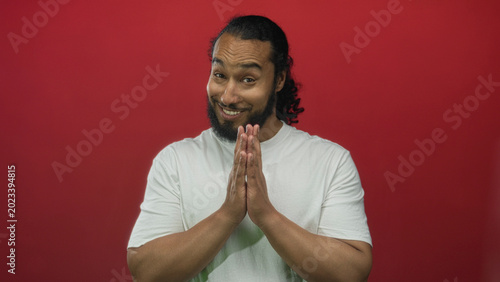 Photos Smiling man in white t shirt with hands pressed together in prayer gesture in red studio; playful plea
