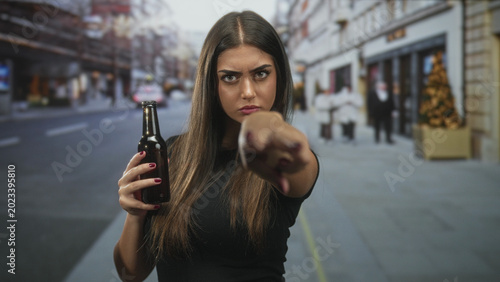 Foto Woman holding beer bottle points finger to camera on a city street with storefronts and christmas tree, visible fingernail and stern gaze; defiance anger