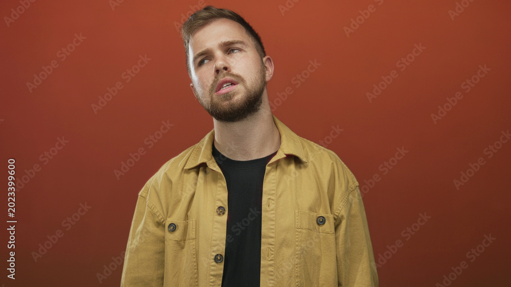 Obraz premium Man tilting head with visible face and beard, wearing yellow jacket against orange studio backdrop; pensive.