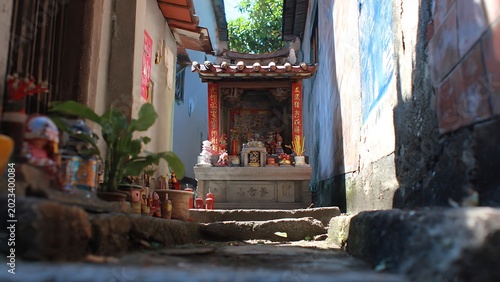 A beautifully adorned traditional outdoor shrine nestled within a narrow, sunlit alleyway, showcasing a peaceful corner of local cultural heritage and spiritual devotion