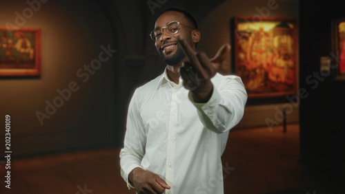 Schilderij op canvas Man points finger forward with visible hand in gallery, smiling while wearing glasses and a white shirt; curiosity and conversation