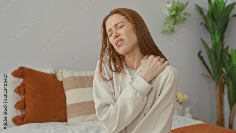 Naklejka premium Woman holding shoulder wincing on bed in building bedroom, eyes closed and hand on shoulder; discomfort recovery.