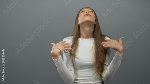 Photography Young hispanic woman shrugging with raised hands and pursed lips, wearing white shirt in gray studio; annoyance disbelief