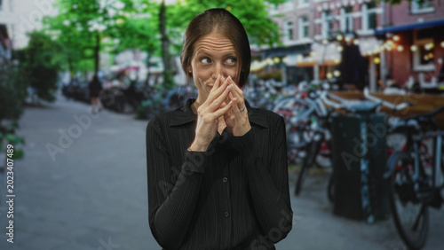 Foto Woman covering mouth with hands on busy city street lined with bicycles and cafe terraces while glancing sideways; shyness curiosity intrigue