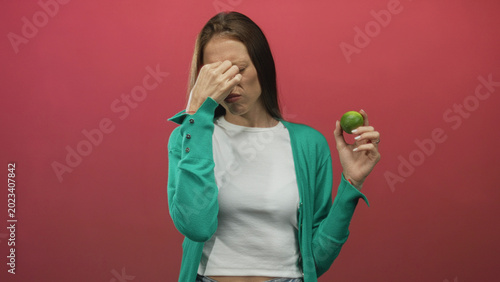 Принти на полотні Young woman holding lime pinching nose, wearing green cardigan and white top, in pink studio; disgust aversion