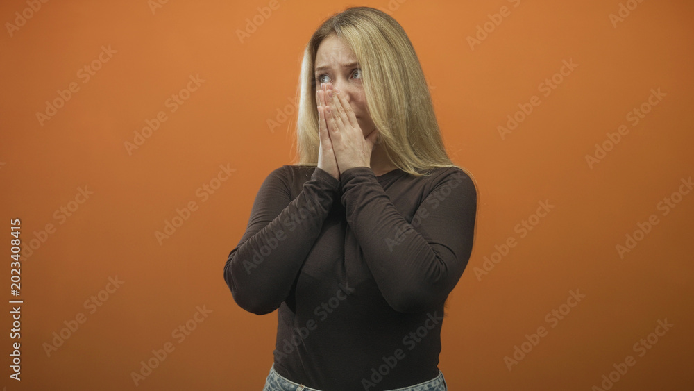 Fototapeta premium Woman biting nails, fingers to mouth, clutching hands at chest, wearing dark top and jeans, standing before orange studio backdrop; anxiety pleading.