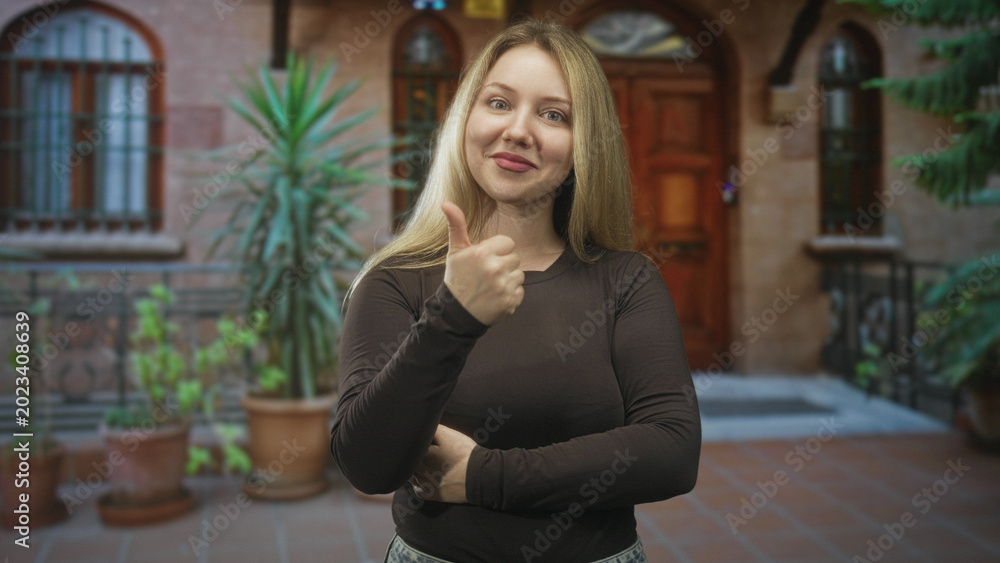 Fototapeta premium Woman showing thumbs up with right hand at building entrance with potted plants and wooden door visible; friendly confidence.