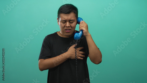Schilderij op canvas Young man holding blue handset to ear while clutching his chest in teal studio; heartache longing yearning
