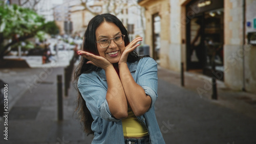 Fotografía Young hispanic woman wearing glasses and denim jacket framing her face with hands and puckered lips on a city street storefront; playful charm