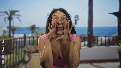Tableau sur toile Woman cups hands to shout on building balcony, mouth open, wearing pink top, glasses and denim jeans; playful excitement