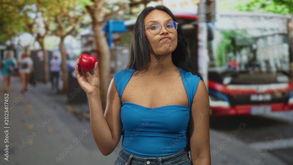 Fototapeta premium Woman holding red heart prop on a busy city street beside a parked bus and pedestrians; health awareness playful.