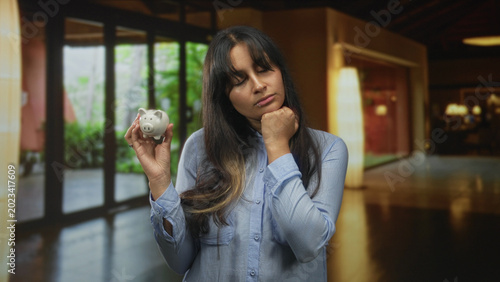 Fényképezés Hispanic woman holding piggy bank, hand on chin and eyes closed in hotel building lobby; financial worry