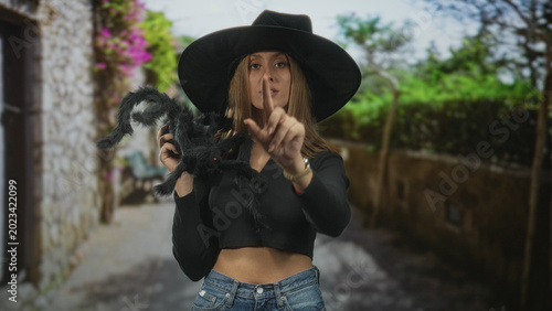 Fotografi Woman in witch hat holds a large black spider prop with one hand, points her finger directly at the viewer while showing a bare midriff in a crop top and jeans on a street; mischief