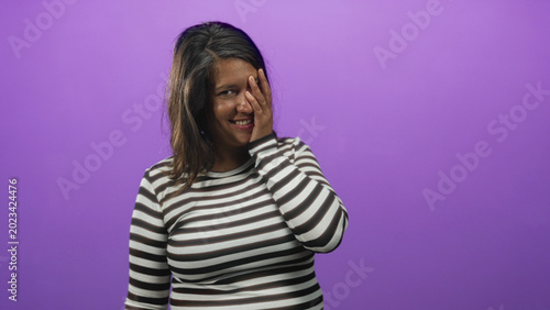 Photos Woman covers face with hand while smiling and tilting her head in a purple studio with a seamless backdrop wearing a striped shirt; bashful joy