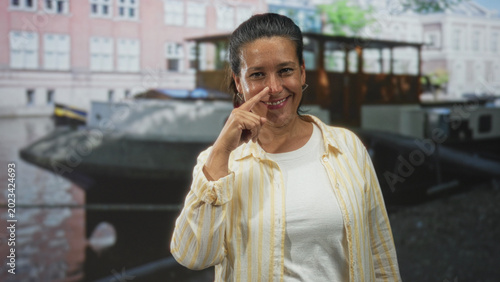 Fotografía Woman points finger to nose while smiling at camera wearing yellow striped shirt and white tee in studio with printed canal backdrop; playful warmth