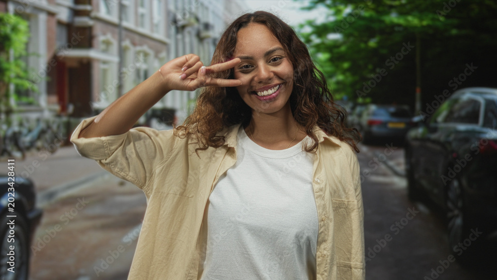 Fototapeta premium Woman making a peace sign with fingers at her eye while smiling in an urban street scene with parked cars and bicycles visible; playful joy.