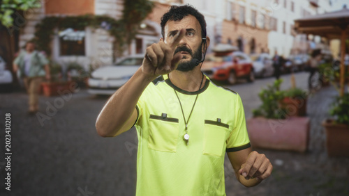 Photos Man referee in neon yellow shirt pointing index finger with whistle on necklace and earpiece, beard visible in street; authority control focus