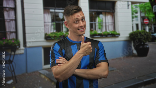 Fotografía Young blond handsome man smiling with crossed arms and thumbs up gesture outside a city building on cobblestone street; confidence camaraderie
