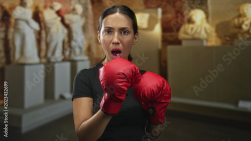 Bild auf Leinwand Woman wearing red boxing gloves with hands raised in a guard stance in a building, mouth open in surprise; surprise strength