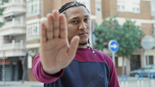 Bild auf Leinwand Man showing outstretched palm stop gesture on urban street with braided hair and sweatshirt; boundary defiance calm