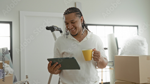 Photos Man holding tablet and yellow mug, smiling amid moving boxes and ladder in apartment; contentment productivity