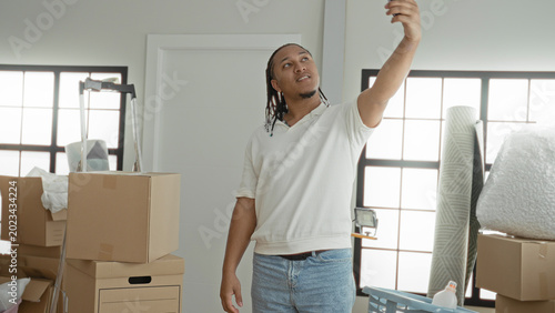 Fotografija Man in white polo holds smartphone aloft taking a selfie among moving boxes inside an apartment building; new home optimism