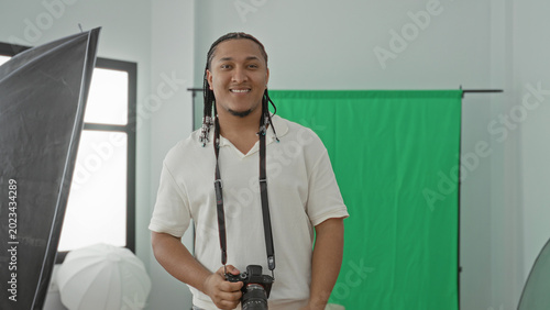 Man aiming camera at lens, holding dslr with both hands in a photo studio with green screen backdrop; creative confidence.