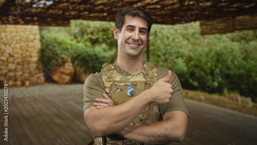 Tableau sur toile Man soldier wearing camouflage vest and dogtags, arms crossed beneath a camo canopy on a forest deck; pride duty