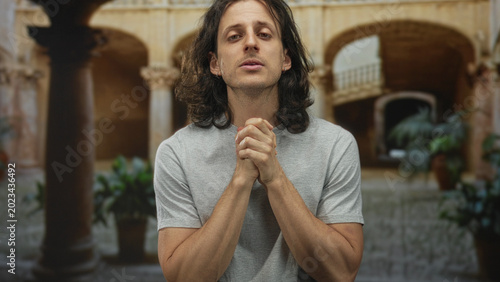 Photography Man with long hair, hands clasped at chest, eyes closed in prayer before old town stone arches and columns of a building courtyard; introspection hope