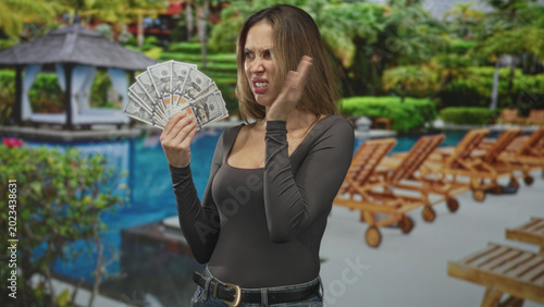 Tableau sur toile Woman holding fan of us dollar bills pinches nose at resort pool building with lounge chairs and gazebo; disgust aversion