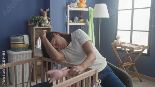 Foto Woman rests head on crib railing, hand over eyes, holding pink toy in baby room with changing table and window; care fatigue