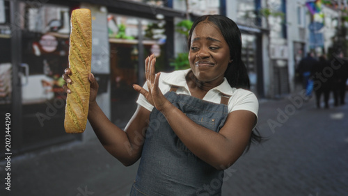 Принти на полотні Woman baker in apron holds a baguette in her left hand and recoils with her right hand in a disgusted grimace on a street; disgust hesitation