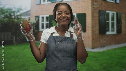 Foto Woman wearing apron and smiling holds a mango with gloved hands in front of brick building and green lawn; homegrown joy