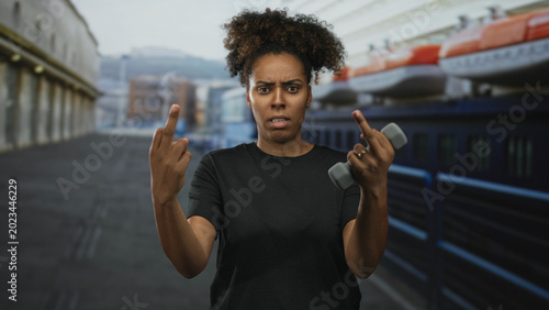Quadro su tela Woman showing middle fingers and holding a dumbbell while standing on cruise ship deck outdoors near lifeboats and railings; anger defiance