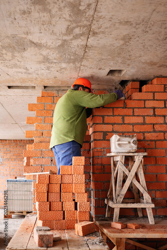 Foto A construction worker in an orange hard hat stands on a wooden platform to build a textured red brick wall up to a bare concrete ceiling in an unfinished room