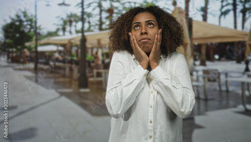 Cuadro en lienzo Young woman cupping cheeks with both hands in a street terrace restaurant near palm trees and outdoor tables; exasperation