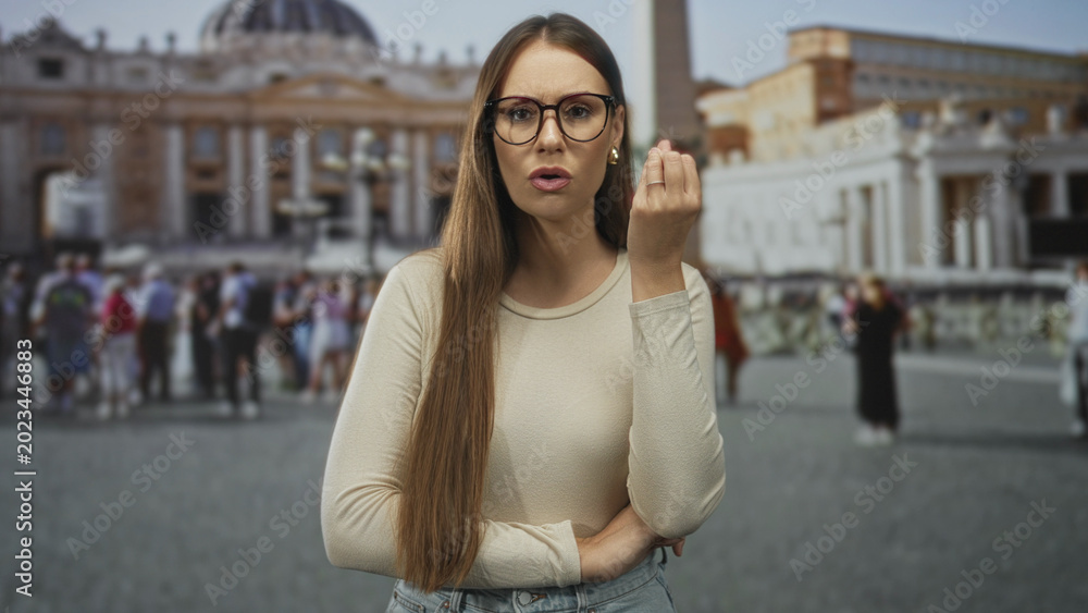 Obraz premium Young woman with glasses holding up hand in pinched fingers gesture in front of building, crowded piazza and basilica visible; impatience tourism.