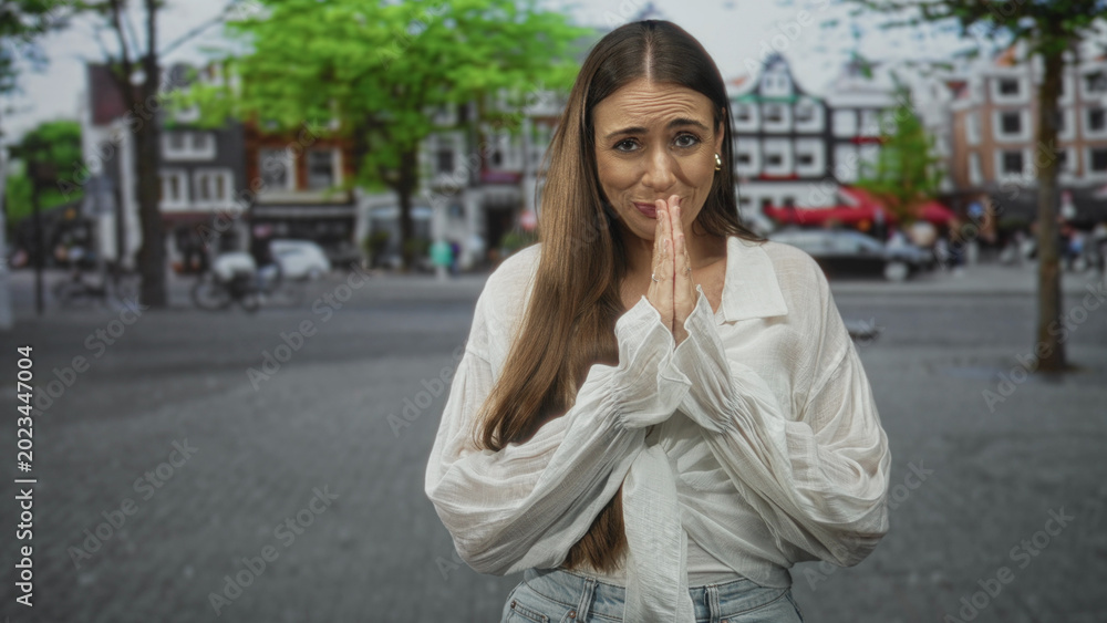 Fototapeta premium Young woman presses hands together near face in pleading gesture on a street lined with cafes and historic buildings; hopeful plea.
