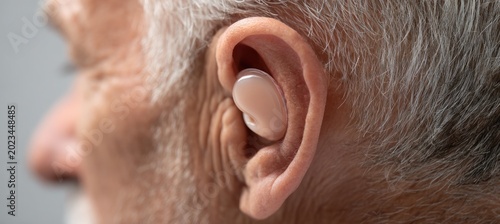 Senior Caucasian man in his 70s with fair skin is wearing a modern beige hearing aid in his ear to improve his auditory sense and daily health care.