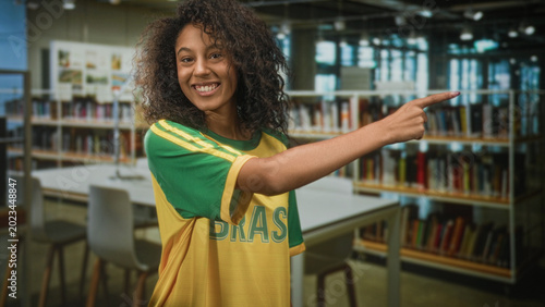 Fotografie Young woman points finger in building library while wearing brazil football tshirt and smiling with curly hair and visible forearm; team spirit joy