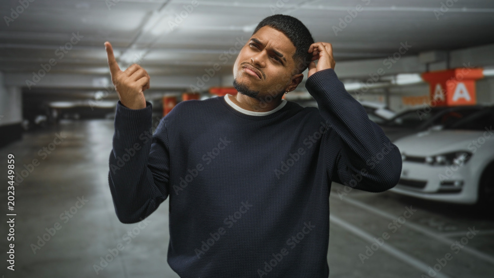 Naklejka premium Hispanic man points finger up and scratches head in an underground parking building beside parked cars and concrete pillars marked a, looking puzzled; confusion, searching.