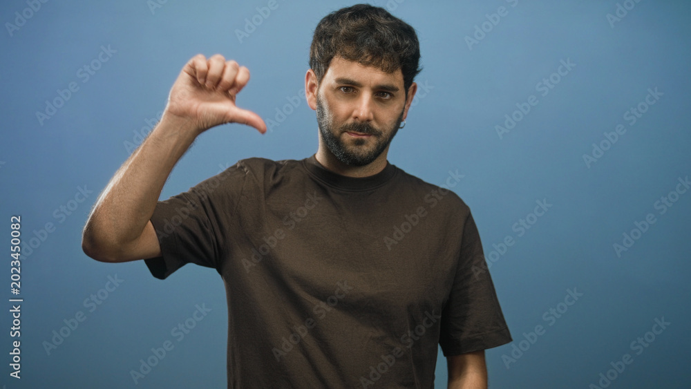 Fototapeta premium Hispanic young man with beard wearing brown t shirt showing thumb down with right hand in studio; disapproval critique.