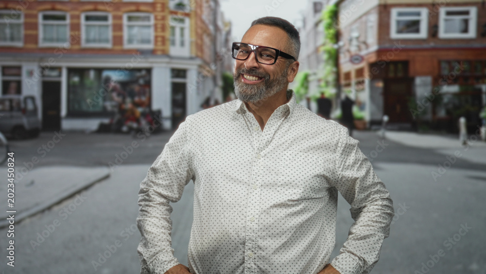 Fototapeta premium Man smiling and pointing hand toward camera on a city street wearing glasses and a white patterned shirt; confidence.