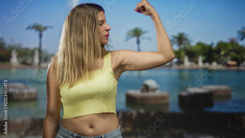 Fotografie Woman flexes right bicep and shows bare midriff wearing yellow crop top and jeans in studio portrait set; confidence strength