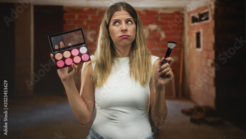 Foto Woman holding makeup palette and brush in an unfinished building, holding brush aloft and frowning at the colors; frustration beauty dilemma