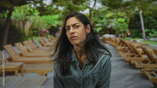 Foto Young woman frowning with visible face and furrowed brow by pool lounge chairs outdoors in denim shirt; uncertainty concern