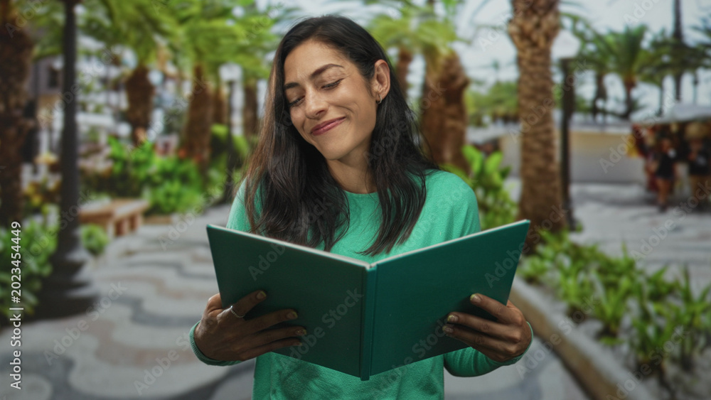 Fototapeta premium Woman reading green book and smiling while holding it on a sunny street lined with palm trees, wearing green sweater; contentment.