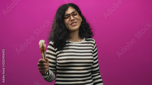 Foto Young arab brunette woman wearing glasses holding makeup brushes in one hand against a pink backdrop in studio, smiling and posing; playful beauty creativity